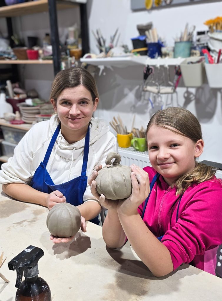 Uśmiechnięta mama z córką trzymające ulepione z gliny dynie podczas warsztatów w pracowni ceramicznej.